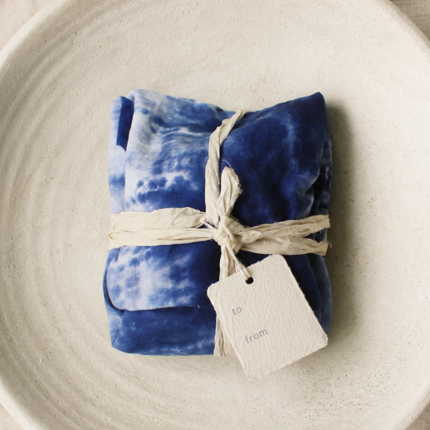 A folded blue and white ice dyed fabric item wrapped in sari ribbon with a small paper tag, placed on a large white ceramic plate over a beige fabric surface.