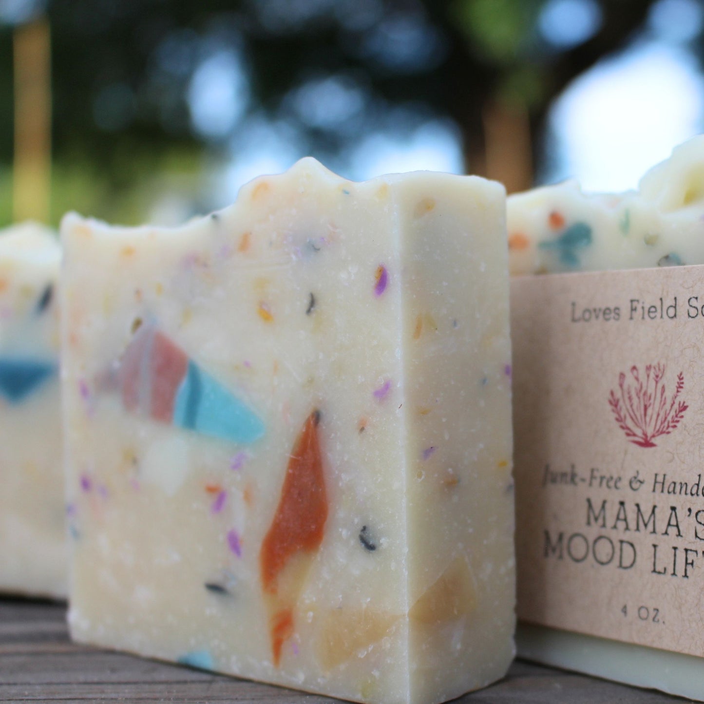 Bar of soap with colorful specks next to a wrapped soap bar that reads 'Mama's Mood Life' on a wooden surface.