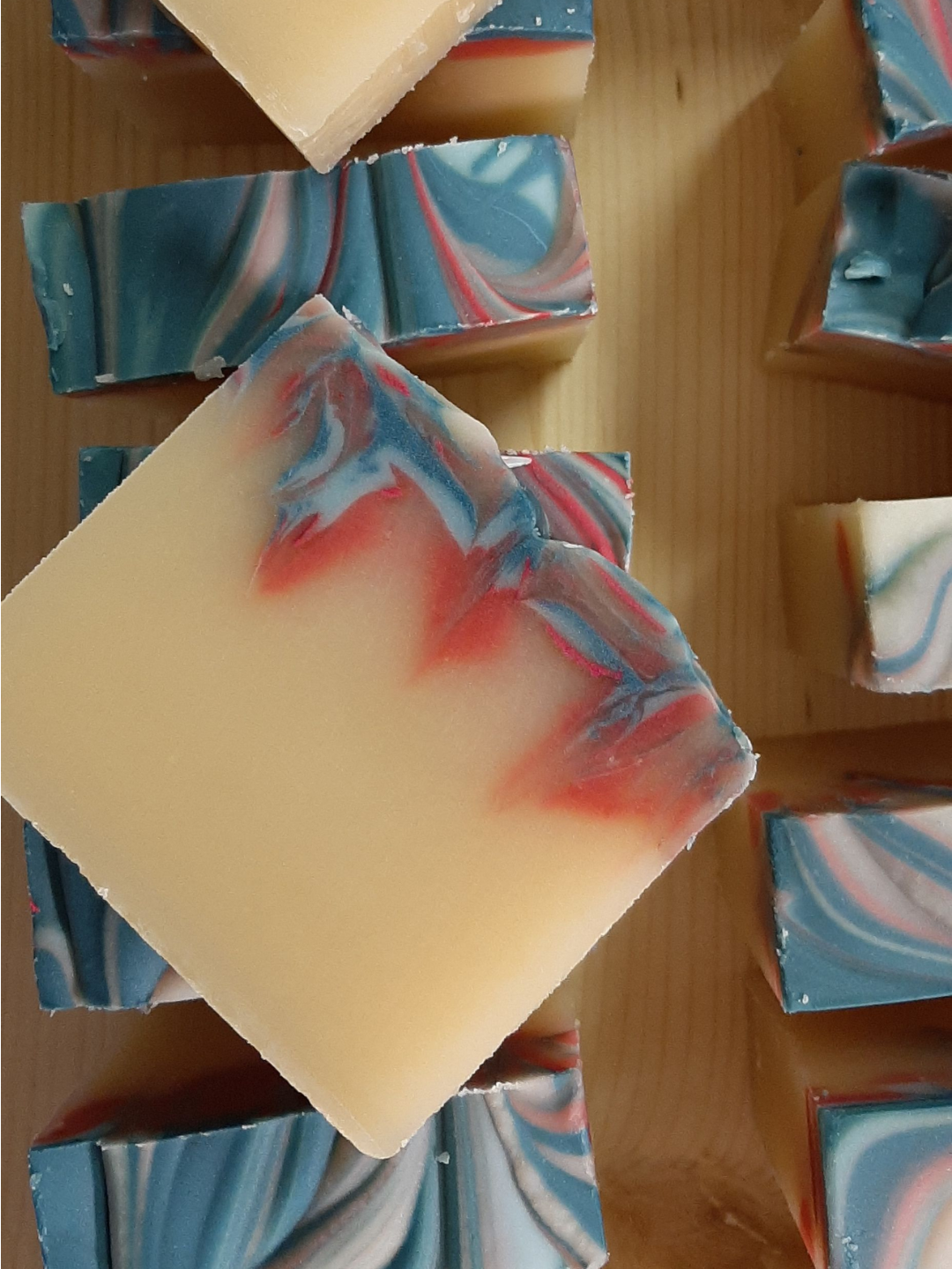 A close-up of handmade Eucalyptus Mint soap bars swirled with blue pink and off white soap bars arranged on a wooden surface, with one large square soap piece in the foreground showing a pale cream base with red and blue swirls.