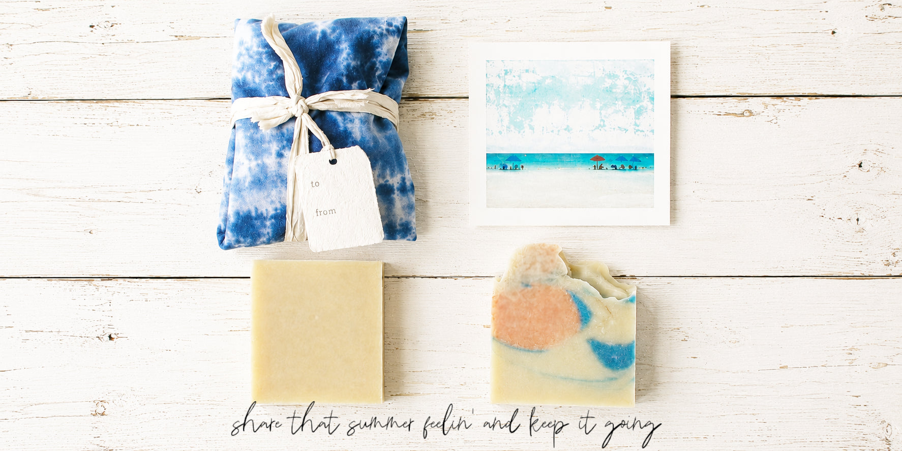 A styled flat-lay of bath products on a white wood background: a wrapped blue-and-white ice-dyed package tied with a string and tag, a small beach-art card, a plain pale yellow bar, and a marbled soap bar with peach, blue, and white tones. Handwritten text at the bottom reads, 'share that summer feelin and keep it going'.