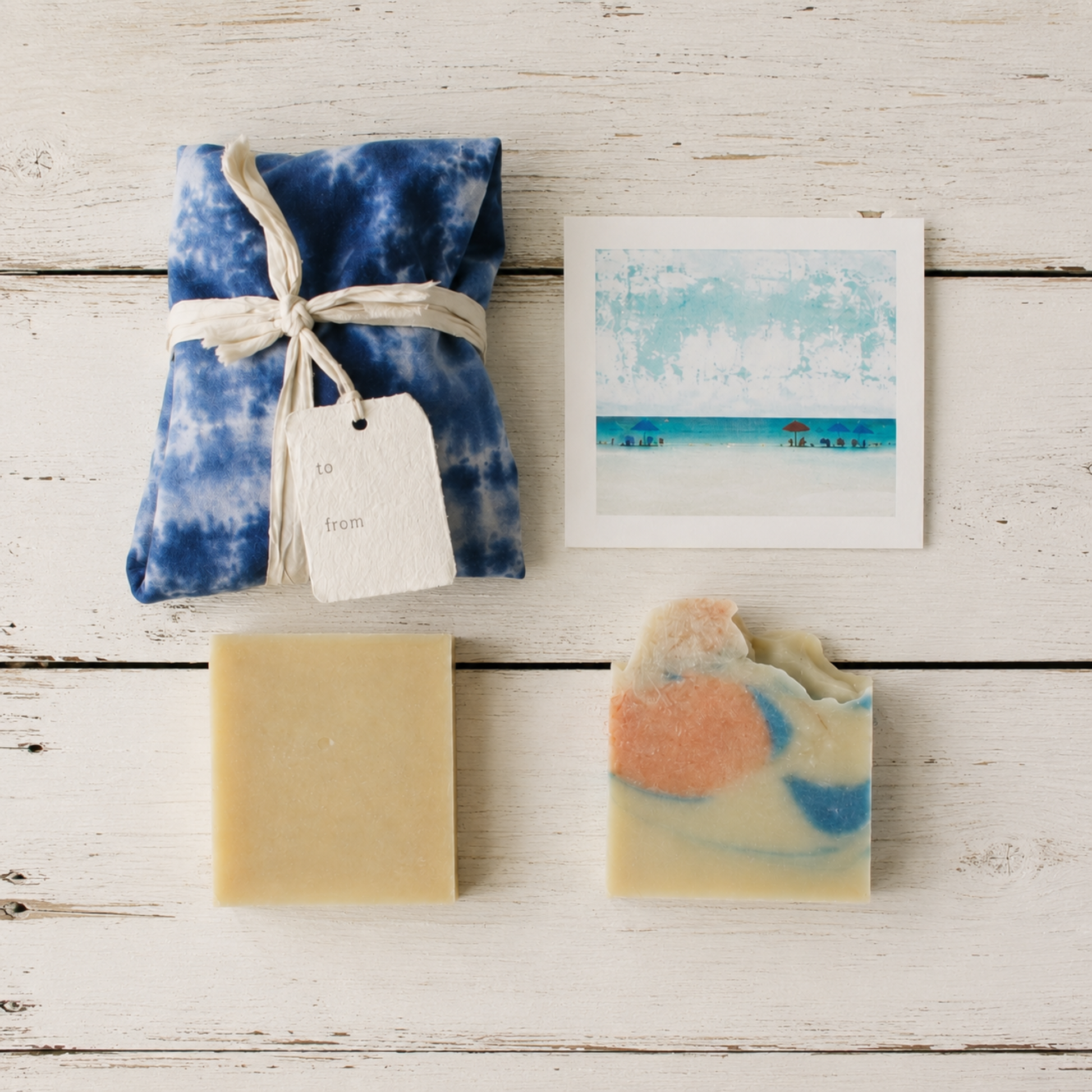 A gift set of handmade soaps and a small art print arranged on a whitewashed wooden surface. The set includes a wrapped blue-and-white ice-dyed fabric package, soap tied with a cream sari ribbon and tag, a plain beige soap bar, a marbled soap with peach, cream, and blue swirls, and a small square beach-themed print.