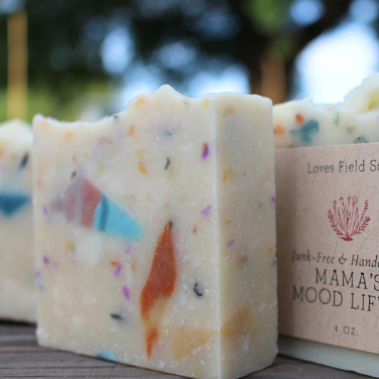 Bar of soap with colorful specks next to a wrapped soap bar that reads  'Mama's Mood Life' on a wooden surface.
