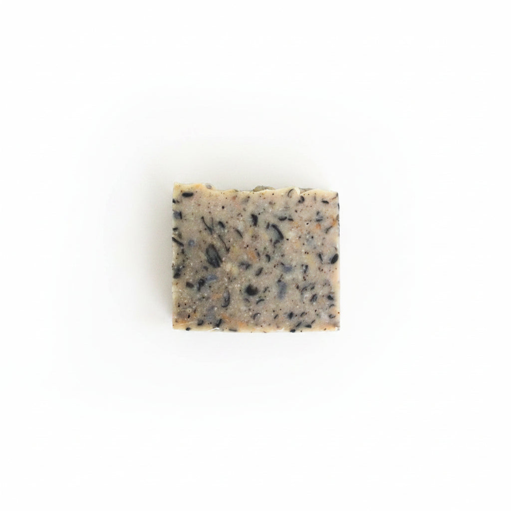 A rectangular bar of handmade soap with black seed inclusions, resting on a textured off-white surface.