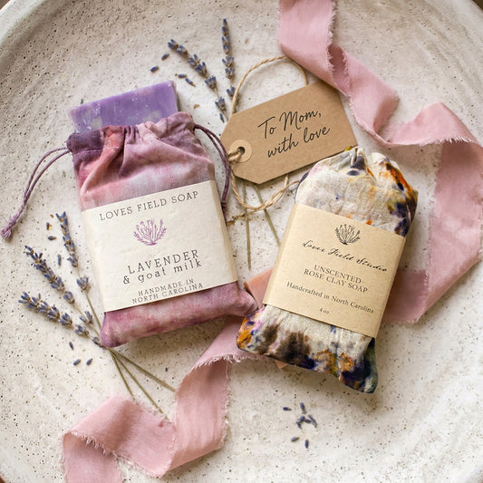 Two soap bars with labels on a textured surface with lavender and pink ribbons.