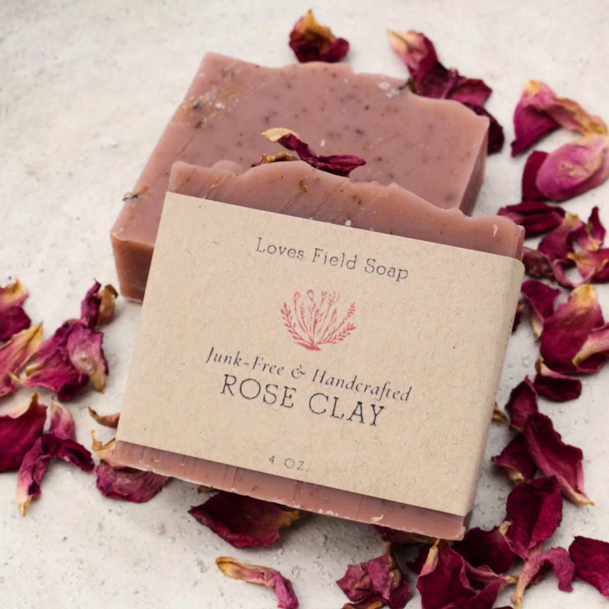 Two bars of handmade rose clay soap by Loves Field Soap, wrapped in a kraft paper label, surrounded by scattered dried rose petals on a light textured surface.
