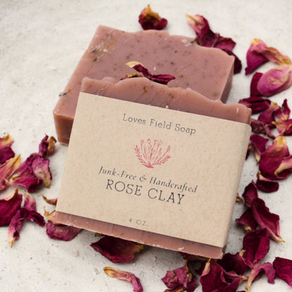 Two bars of handmade rose clay soap by Loves Field Soap, wrapped in a kraft paper label, surrounded by scattered dried rose petals on a light textured surface.